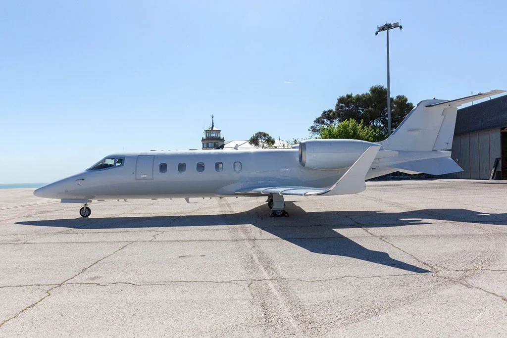 A white Learjet business jet parked on an airport tarmac with a control tower and ocean in the background.