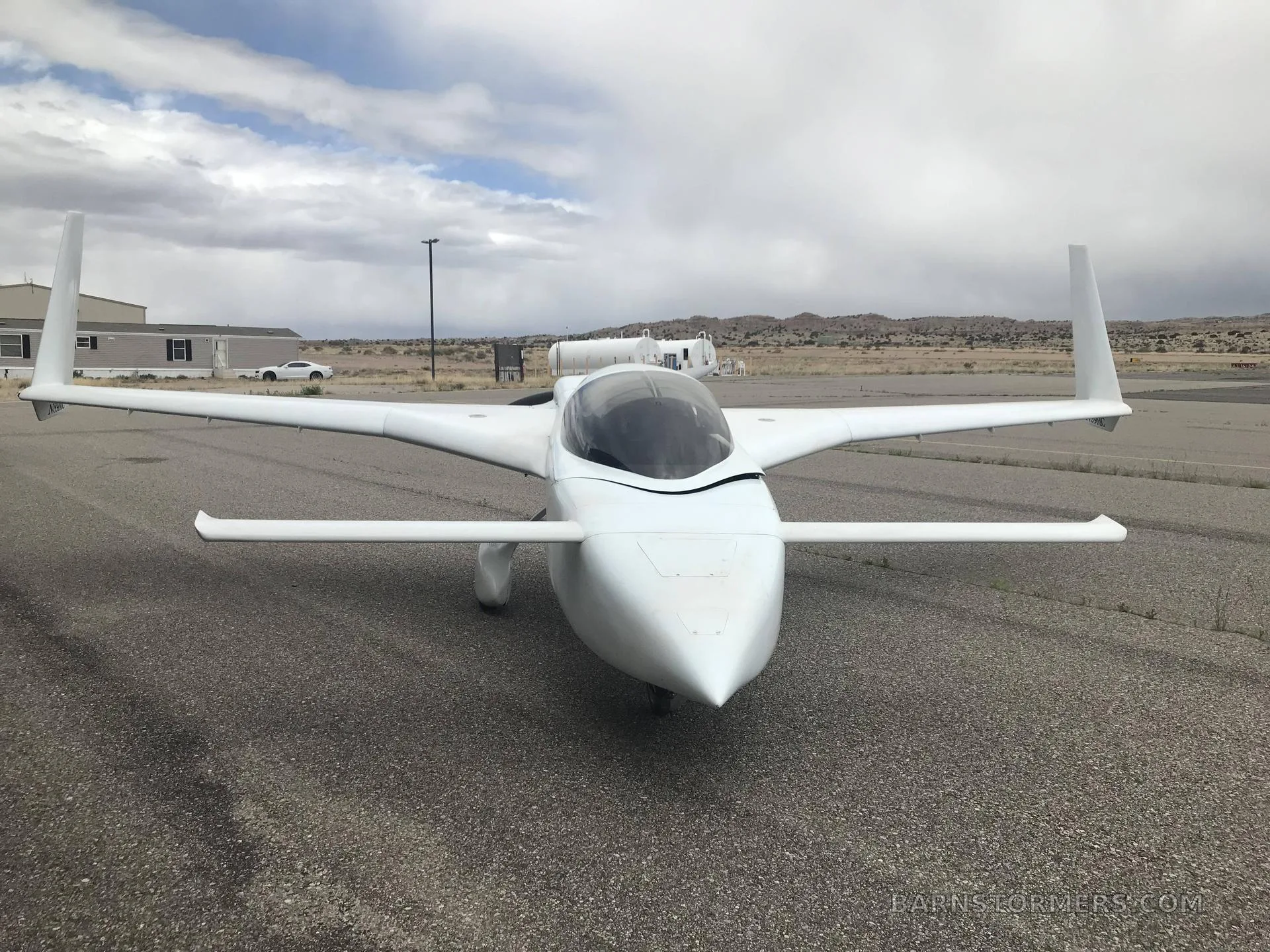 A white, sleek, canard-wing aircraft with twin vertical stabilizers is parked on an asphalt surface under a cloudy sky.