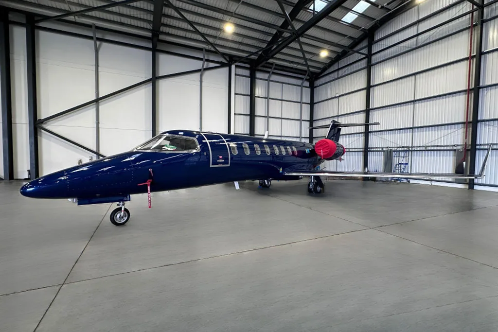 A dark blue private jet with red engine covers parked inside a large hangar, viewed from the front-left.