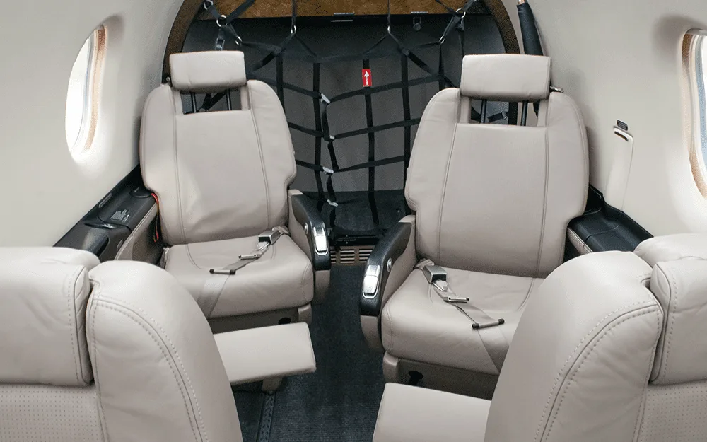 The beige leather interior cabin of a Pilatus PC-12 NG, showing passenger seats and cargo netting in the rear.