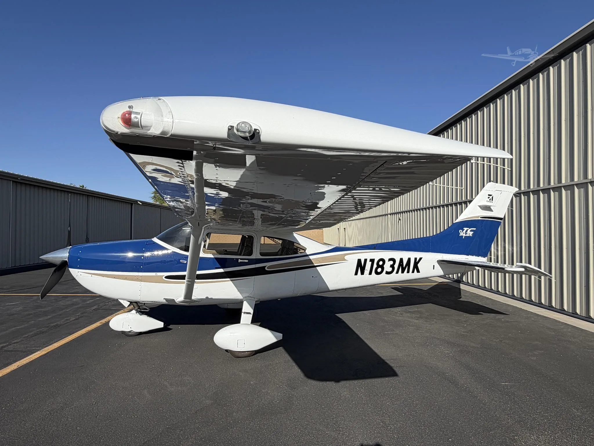 A blue and white Cessna 182 Skylane (N183MK) is parked on an asphalt ramp next to a hangar under a clear blue sky.