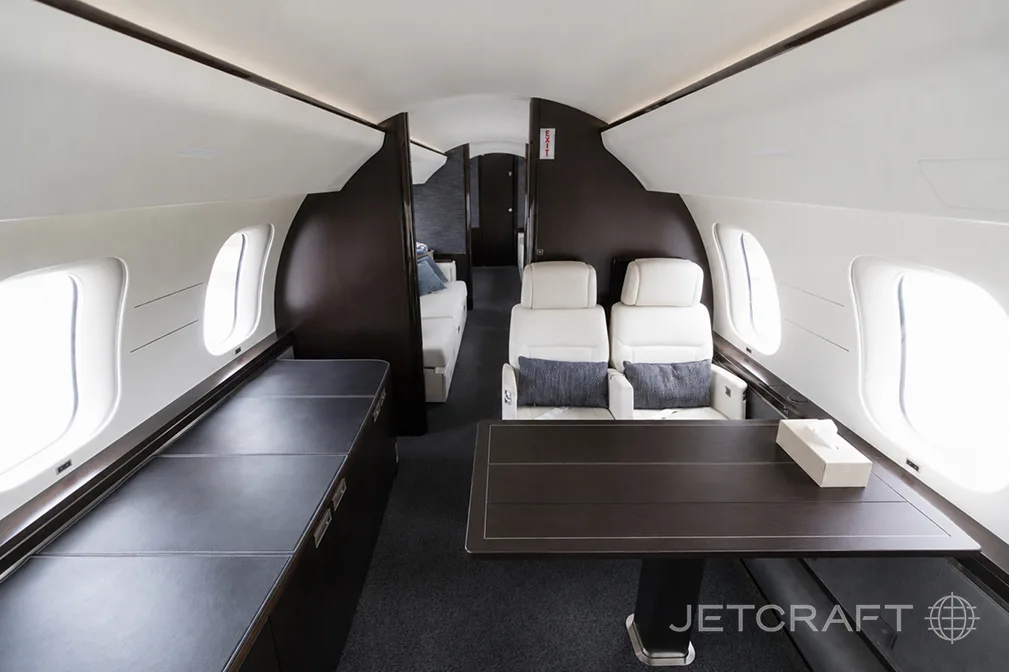A section of the Bombardier Global 7500 cabin with a dark wood table, two white seats, and a sofa in the background.
