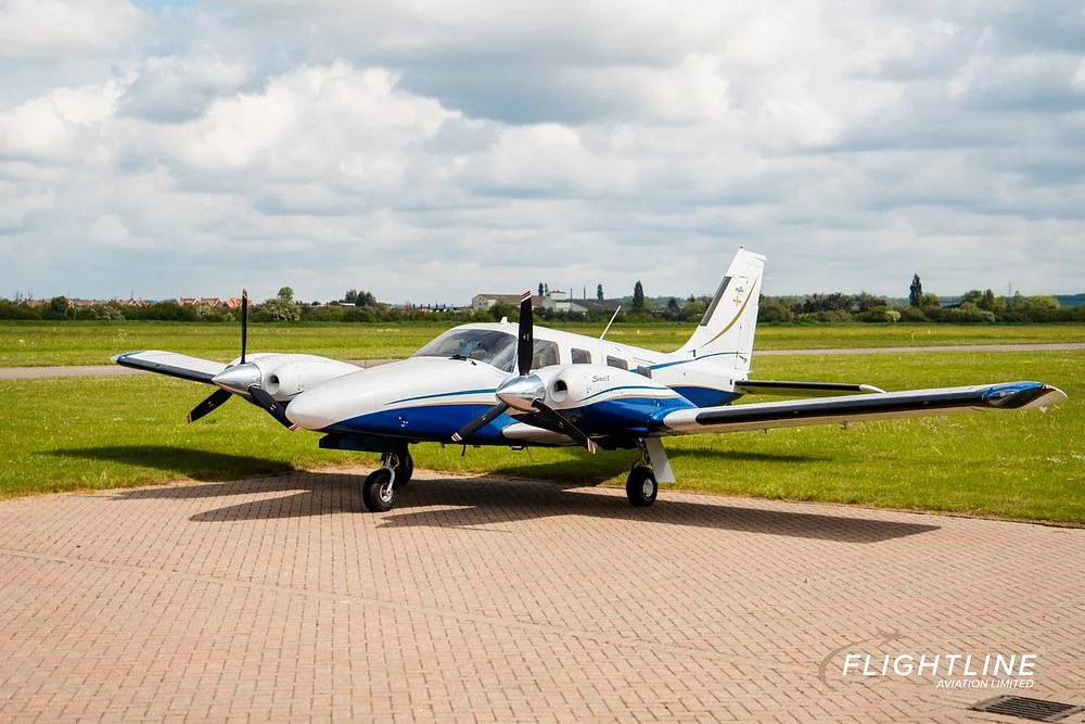 A white and blue twin-engine Piper Seneca aircraft is parked on a tarmac under a cloudy sky.