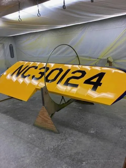 A yellow biplane wing with black registration NC 30124 is propped up in a workshop, possibly during painting or repair.