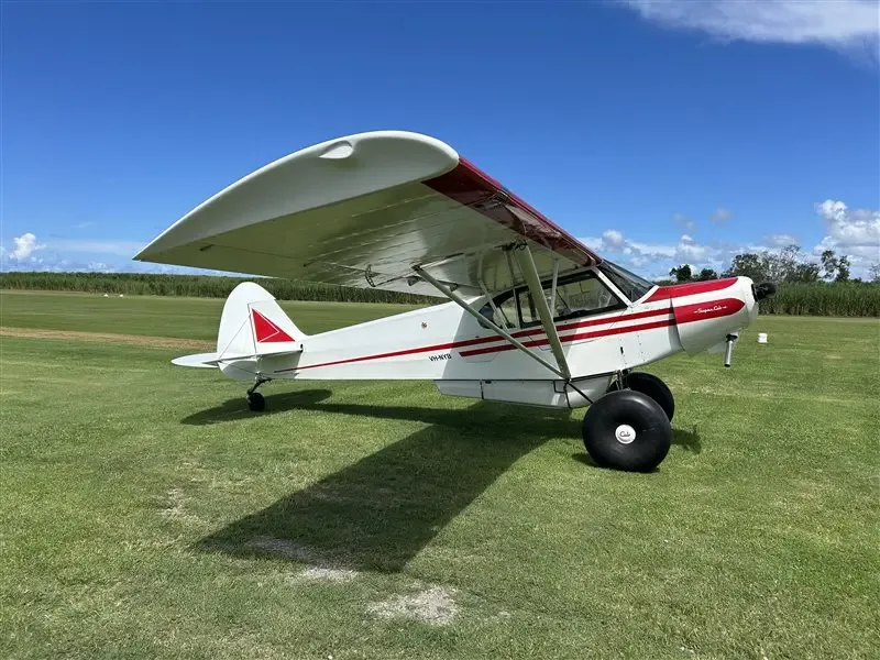 A white and red Super Cub aircraft with large tundra tires parked on a grassy field under a blue sky.
