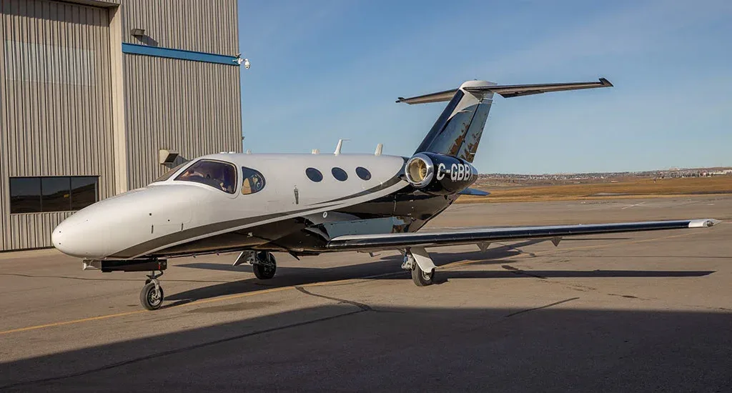 A white and black private jet, registration C-GBBK, parked on an asphalt apron next to a hangar under a clear sky.