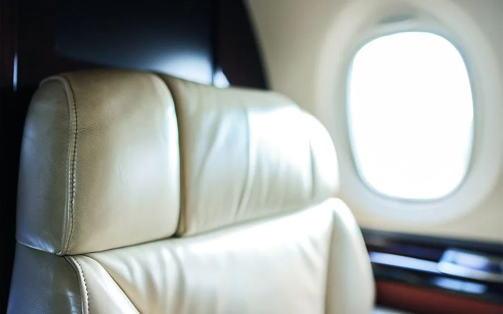 A close-up of a cream-colored leather aircraft seat with a bright oval window in the background.