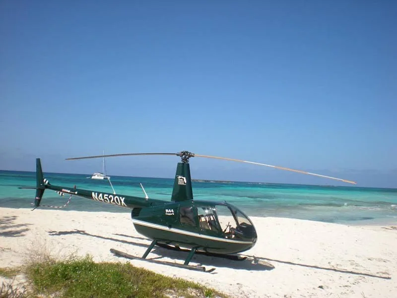A dark green Robinson R44 Clipper II helicopter, registration N4520X, sits on a white sand beach next to turquoise water under a clear blue sky.