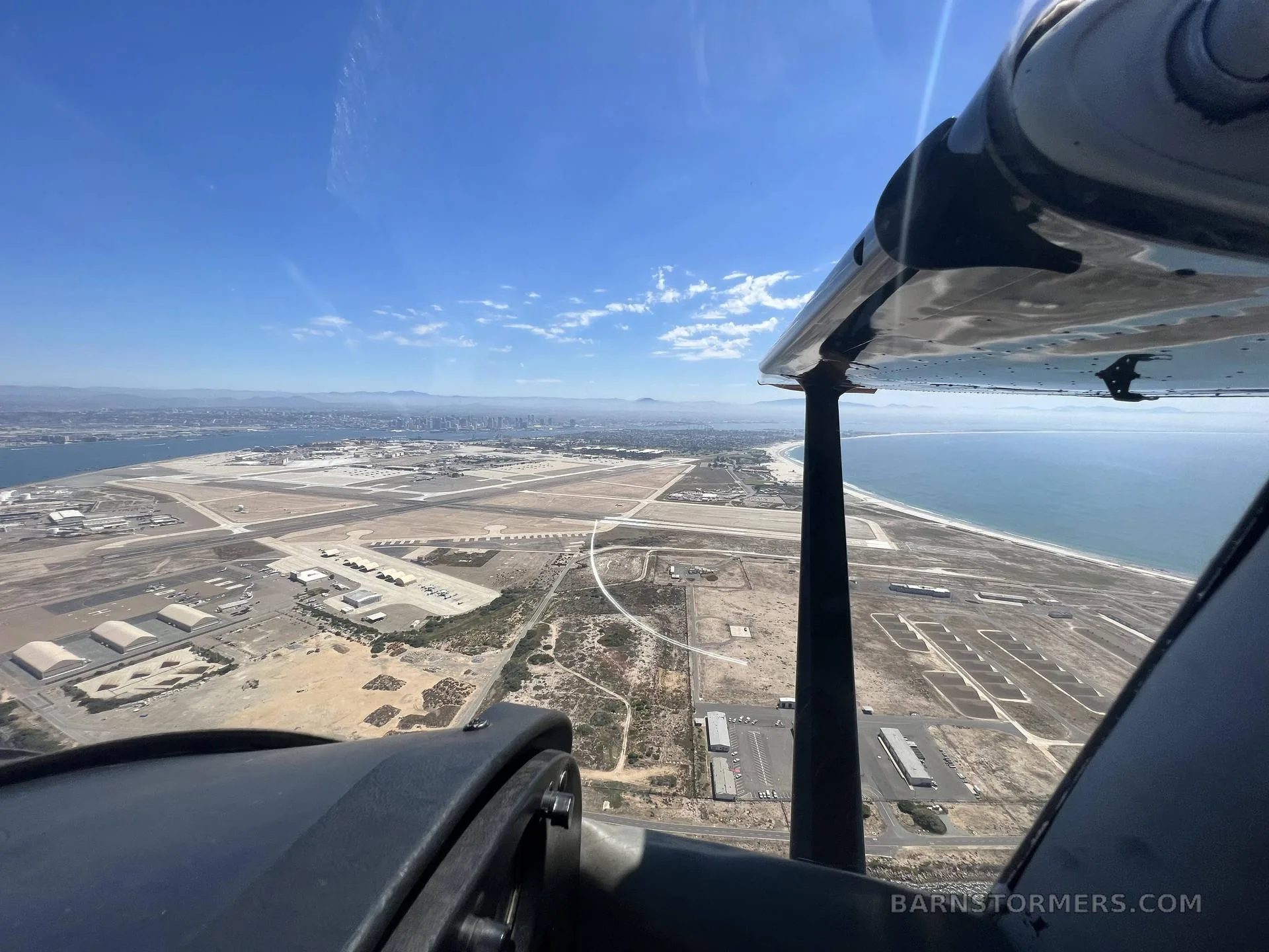 An aerial view from an aircraft showing a large airport, city skyline, and ocean under a clear blue sky.