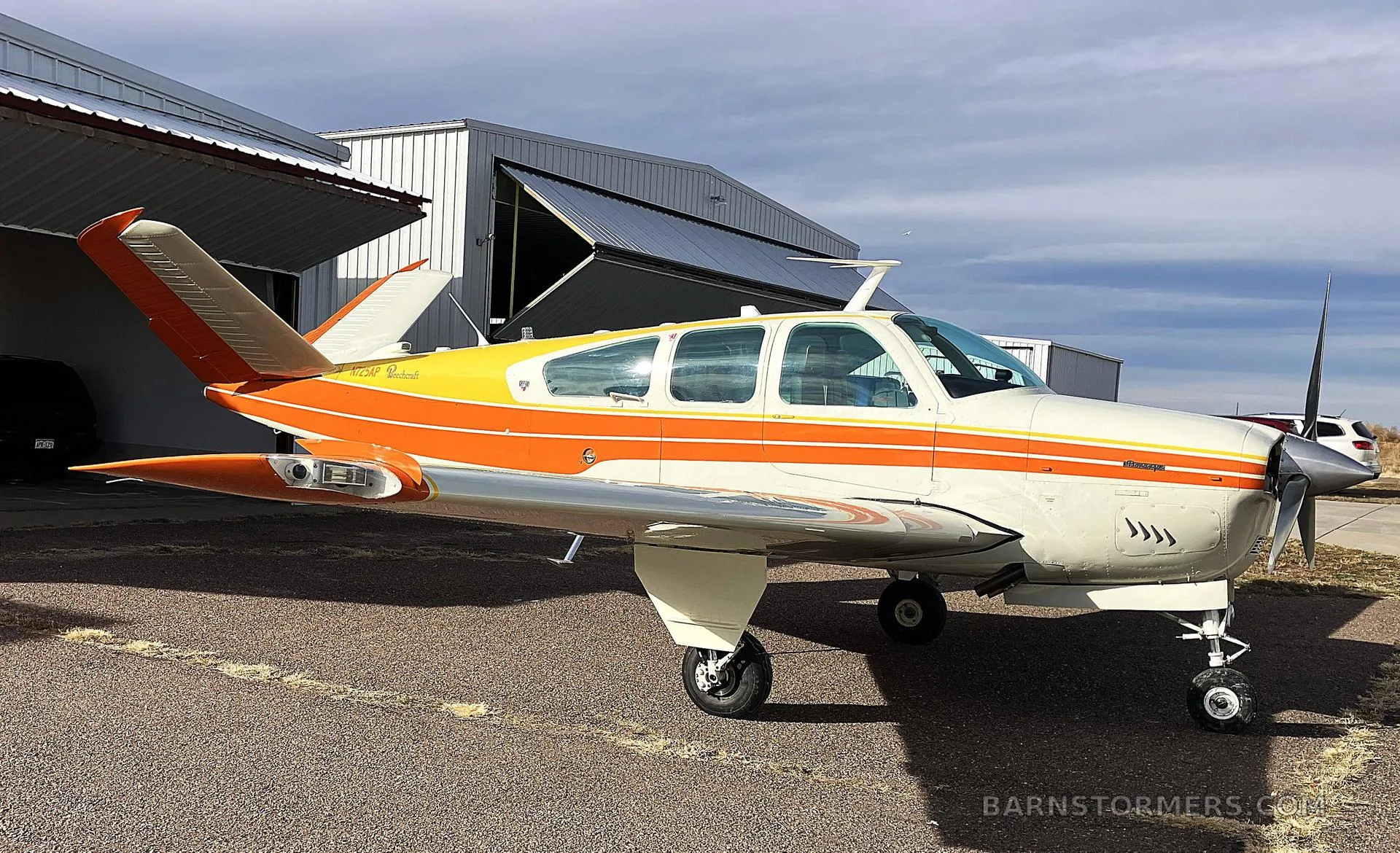 A front-quarter view of a white, orange, and yellow Beechcraft Bonanza N725AP parked on an airfield with an open hangar behind it.