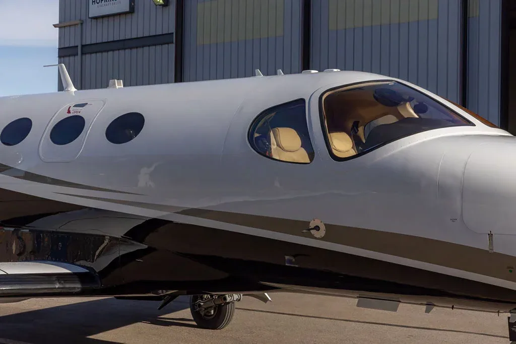 A close-up of the front fuselage and cockpit windows of a white and black private jet, showing the tan interior seats.