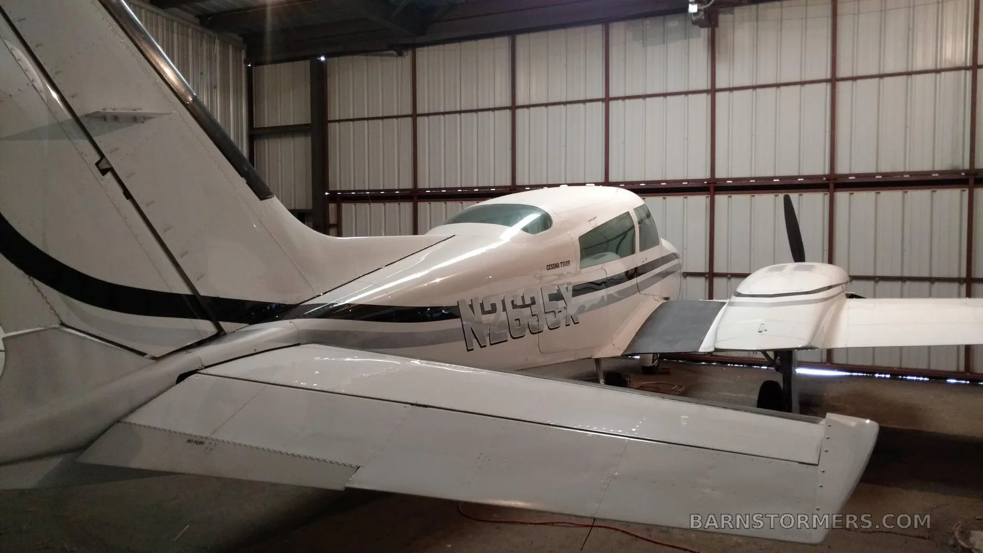 The rear-right view of a white Cessna 310 twin-engine aircraft with black stripes parked inside a metal hangar.