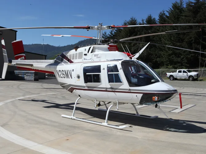 A white Bell 206B JetRanger III helicopter with red and grey stripes is parked on a concrete helipad on a sunny day.