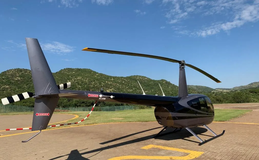 A rear-side view of a grey and black Robinson R44 helicopter on a helipad under a clear blue sky.