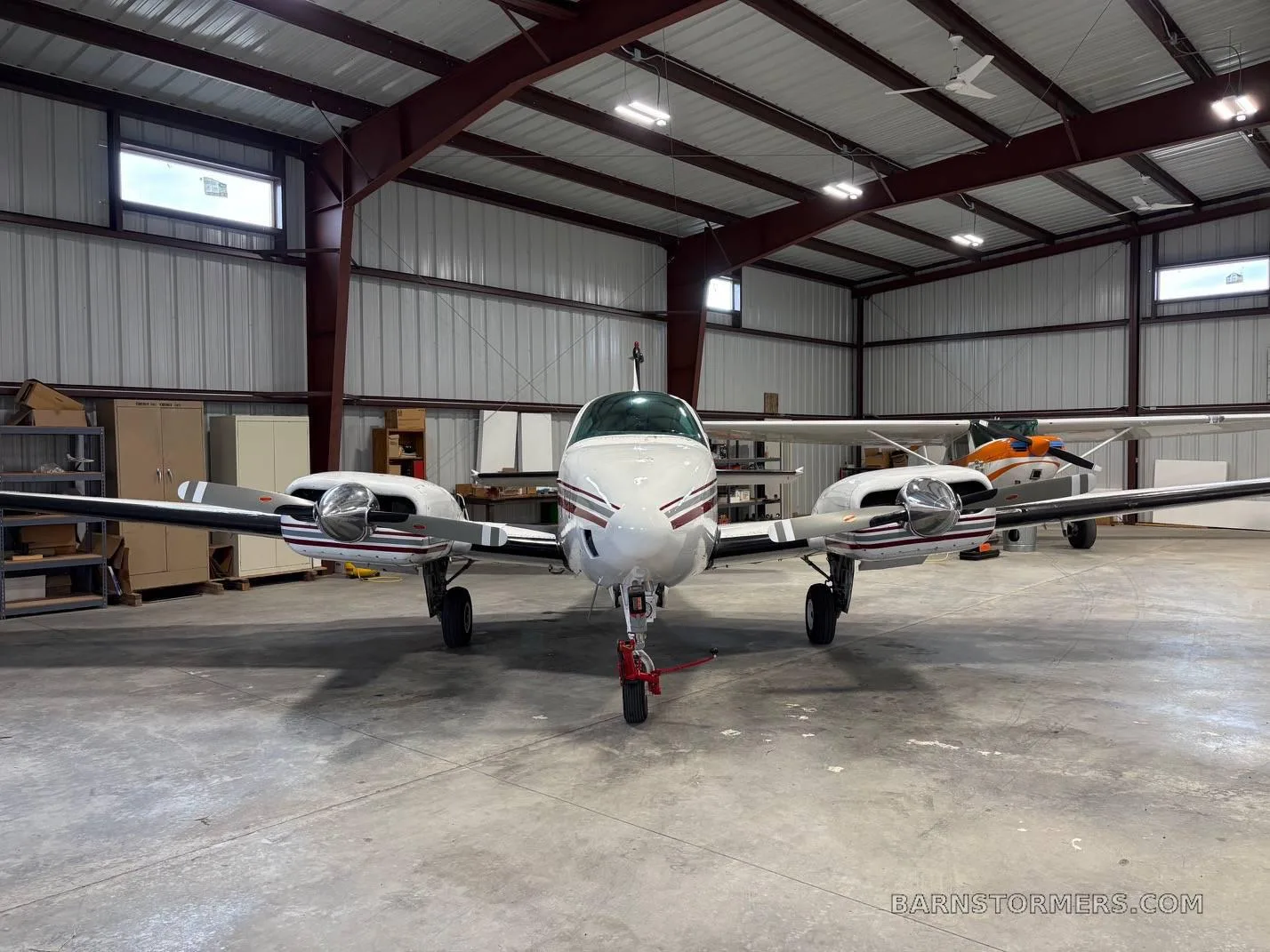A white twin-engine propeller aircraft with red and grey stripes, seen from the front in a hangar.