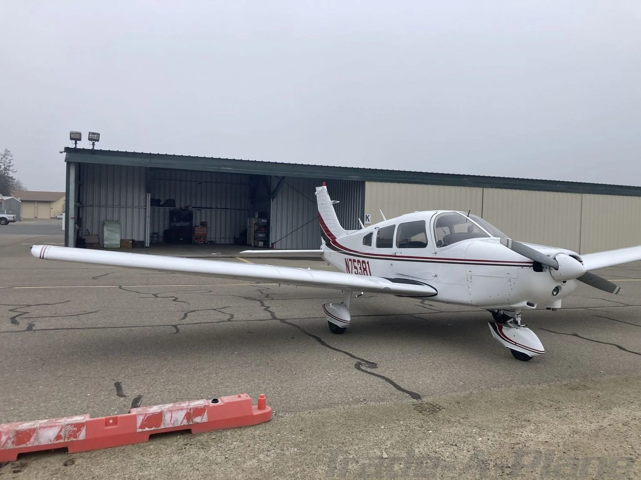 A white Piper PA-28-151 aircraft with red and black stripes parked on an airfield next to an open hangar.
