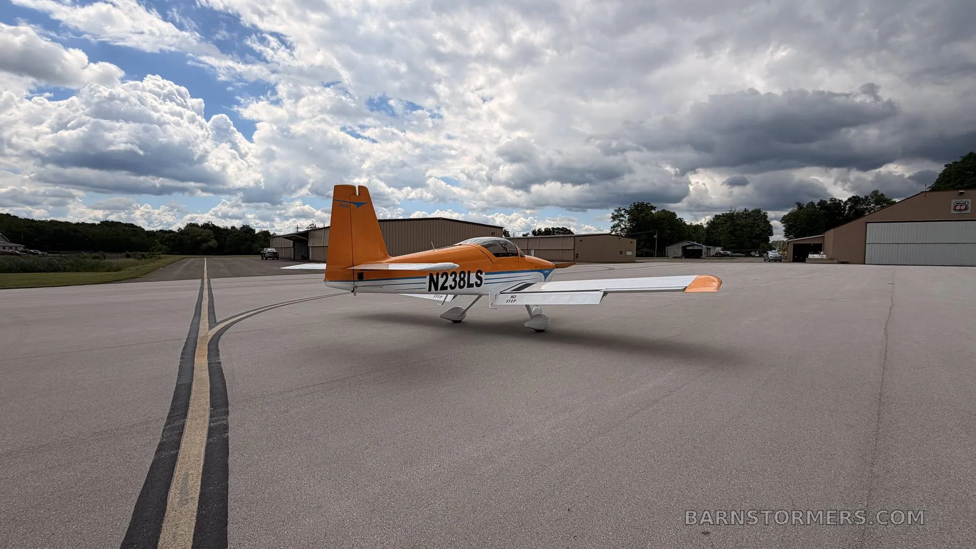 A three-quarter rear view of an orange and white single-engine aircraft with registration N238LS on an airfield, with hangars in the background.