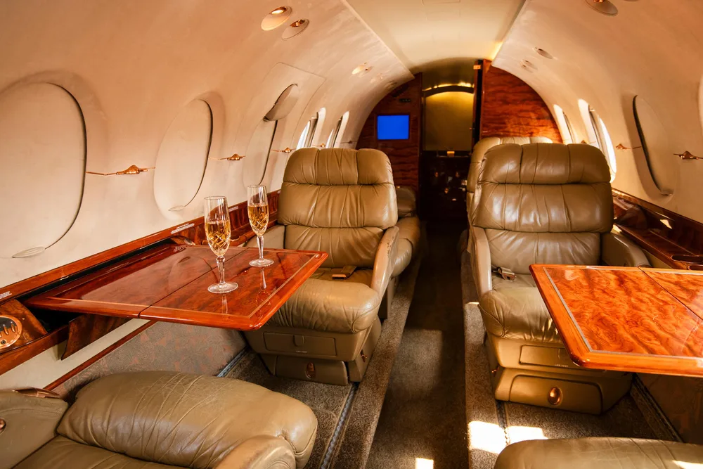 The luxurious interior cabin of a private jet features tan leather seats, polished wood tables, and two champagne flutes.