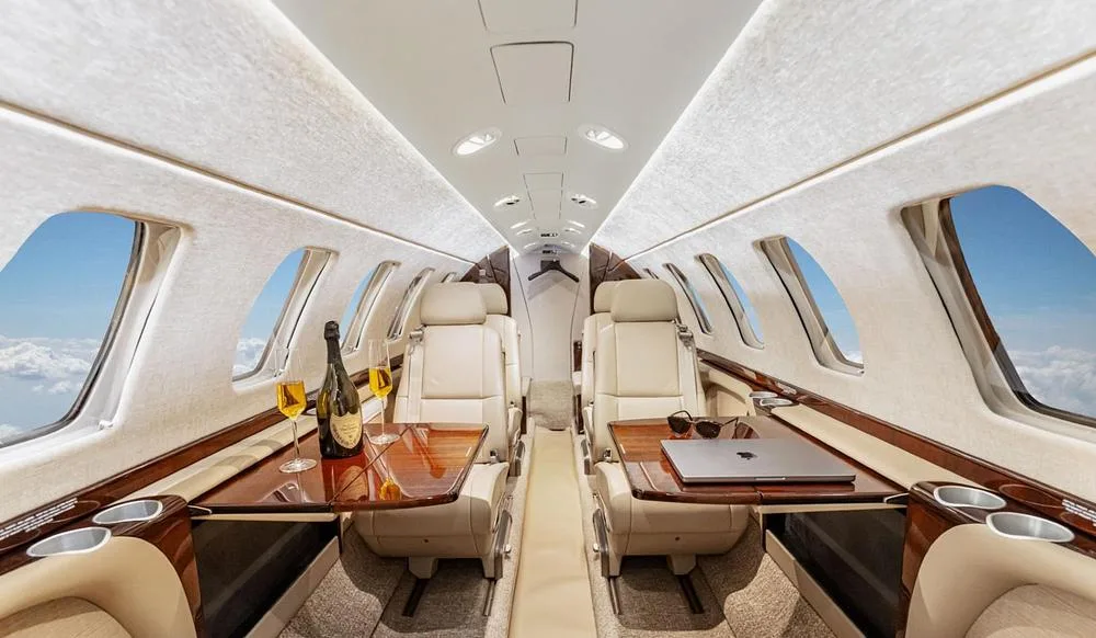 The luxurious interior cabin of a private jet, featuring beige leather seats, polished wooden tables with champagne, and views of clouds from the windows.