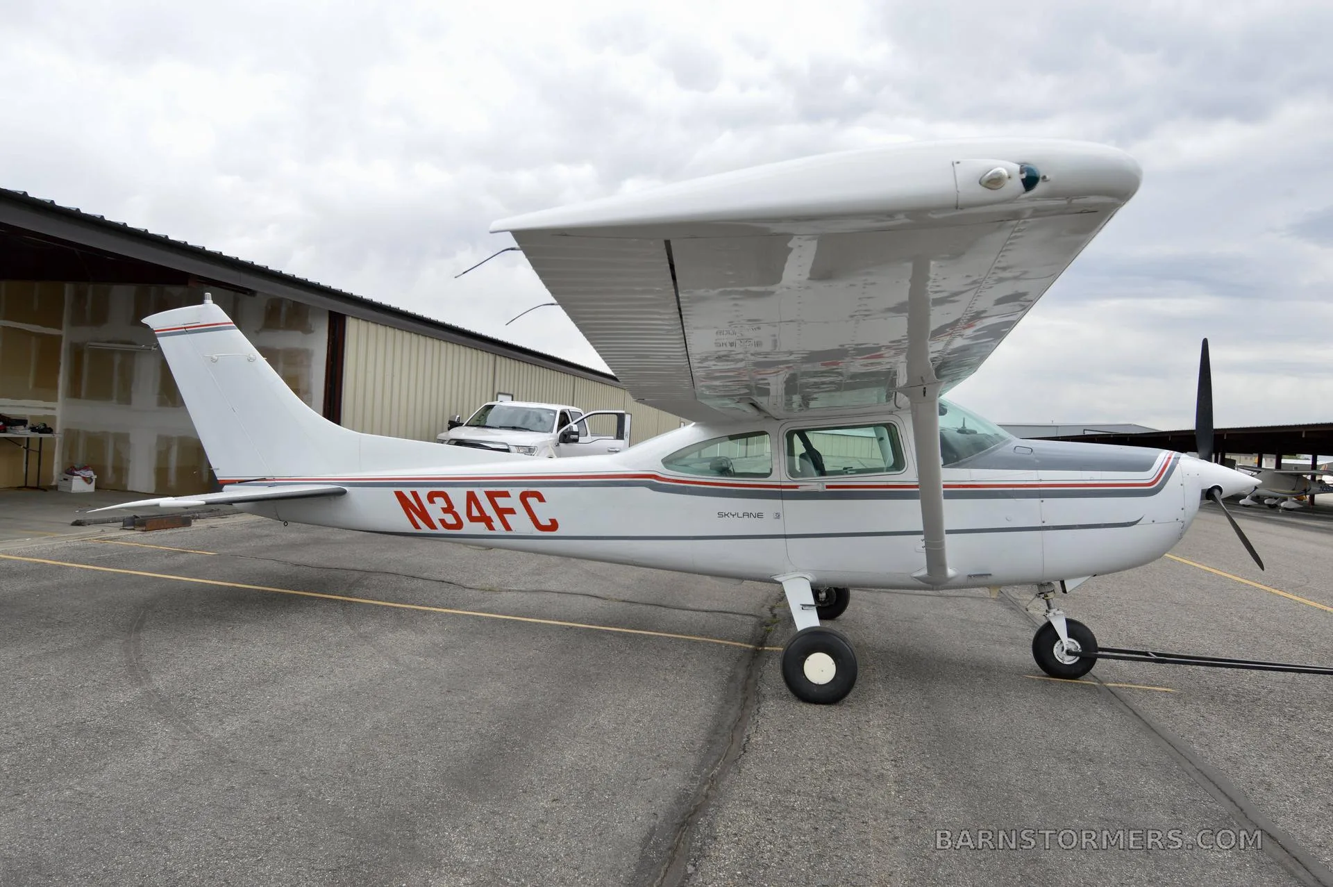 A white Cessna 182 Skylane with red and gray stripes, registration N34FC, parked on an asphalt ramp next to a hangar.
