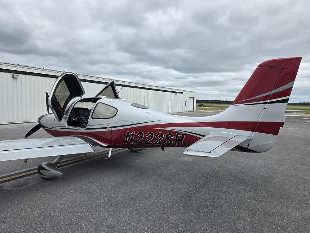 A white, red, and black single-engine aircraft with open doors, viewed from the rear-left on a tarmac.