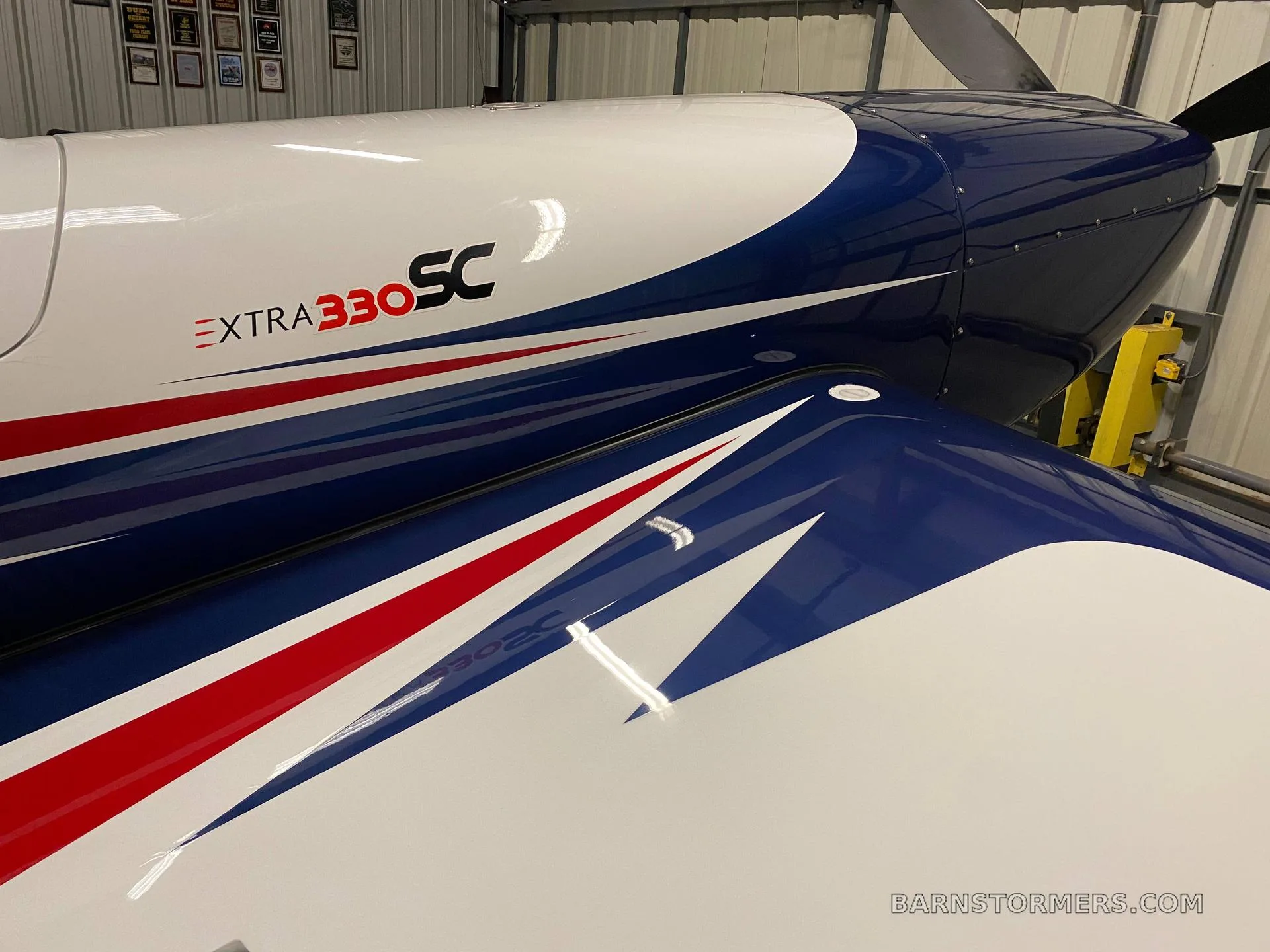 A close-up view of the blue, white, and red striped fuselage and wing of an Extra 330SC aircraft in a hangar.