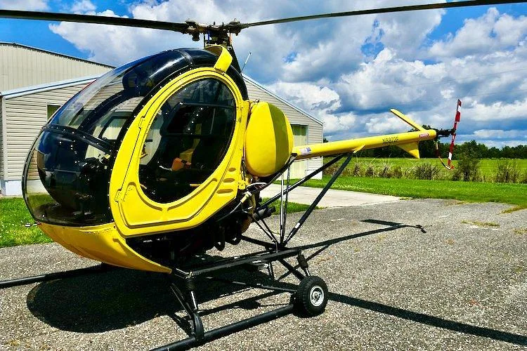 A yellow single-engine helicopter, N8872F, parked on an asphalt pad in front of a hangar under a blue sky with clouds.