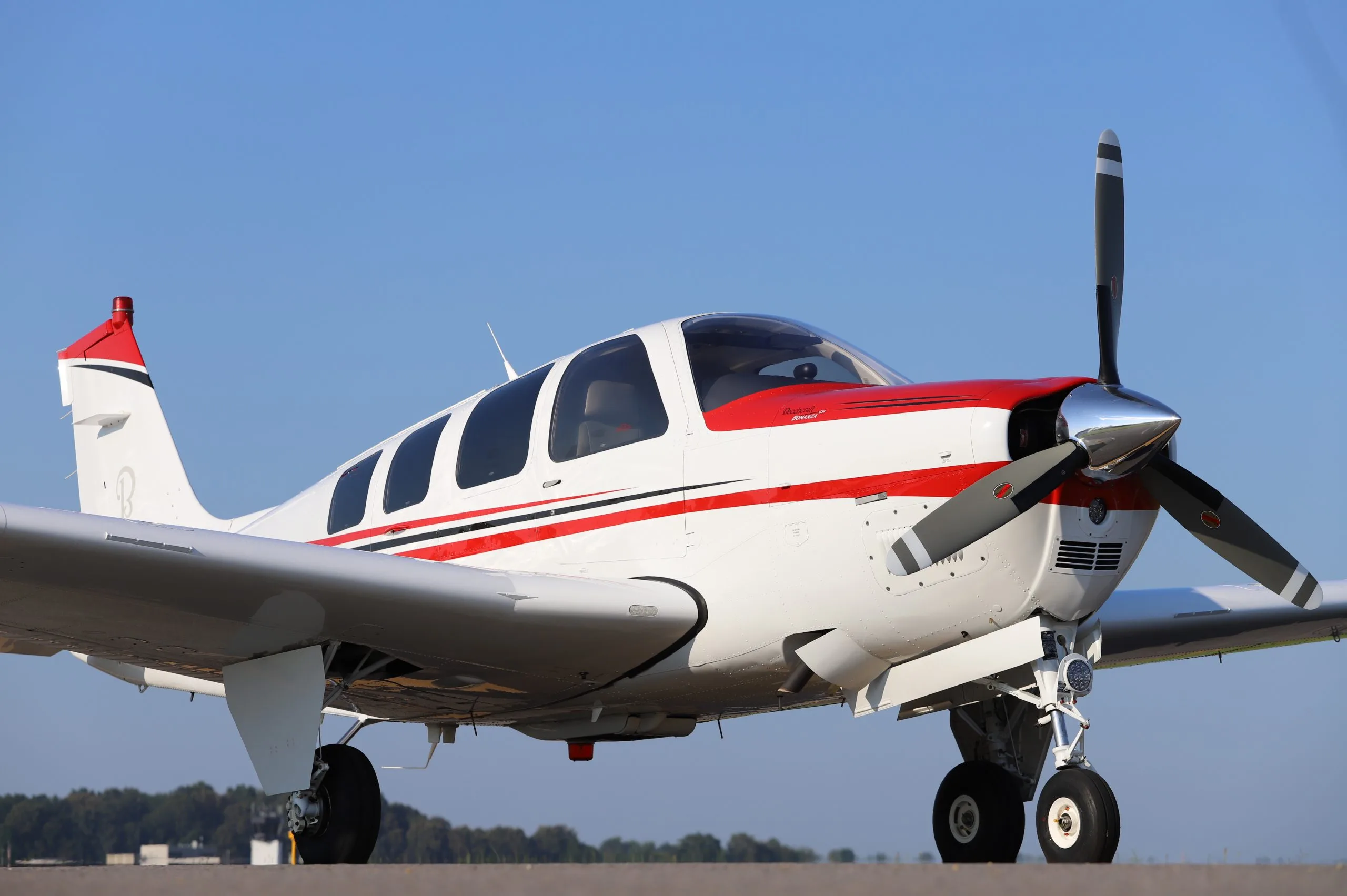 A white Beechcraft Bonanza G36 with red and black stripes is parked on a tarmac under a clear blue sky.