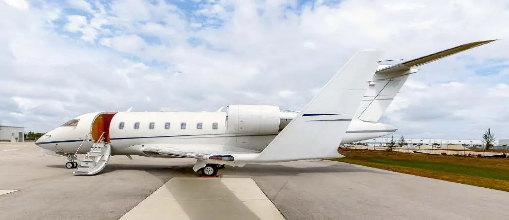 A white business jet, possibly a Bombardier Challenger, with its main door open and stairs extended, parked on an airfield under a cloudy sky.