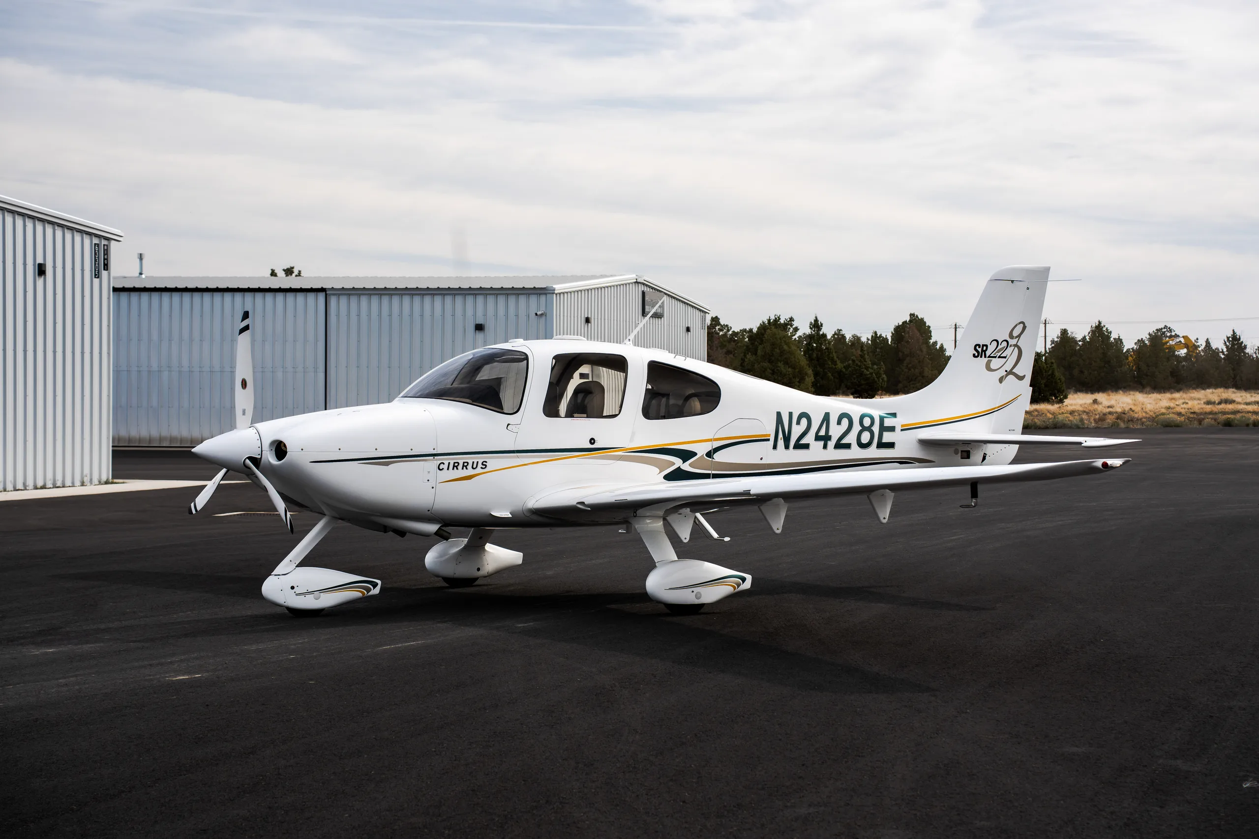 A white Cirrus SR22 aircraft with green and gold stripes, tail number N2428E, parked on an airfield next to hangars.