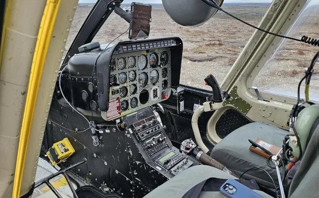 The cockpit of a helicopter featuring an analog instrument panel, cyclic, collective, and a pilot's seat.