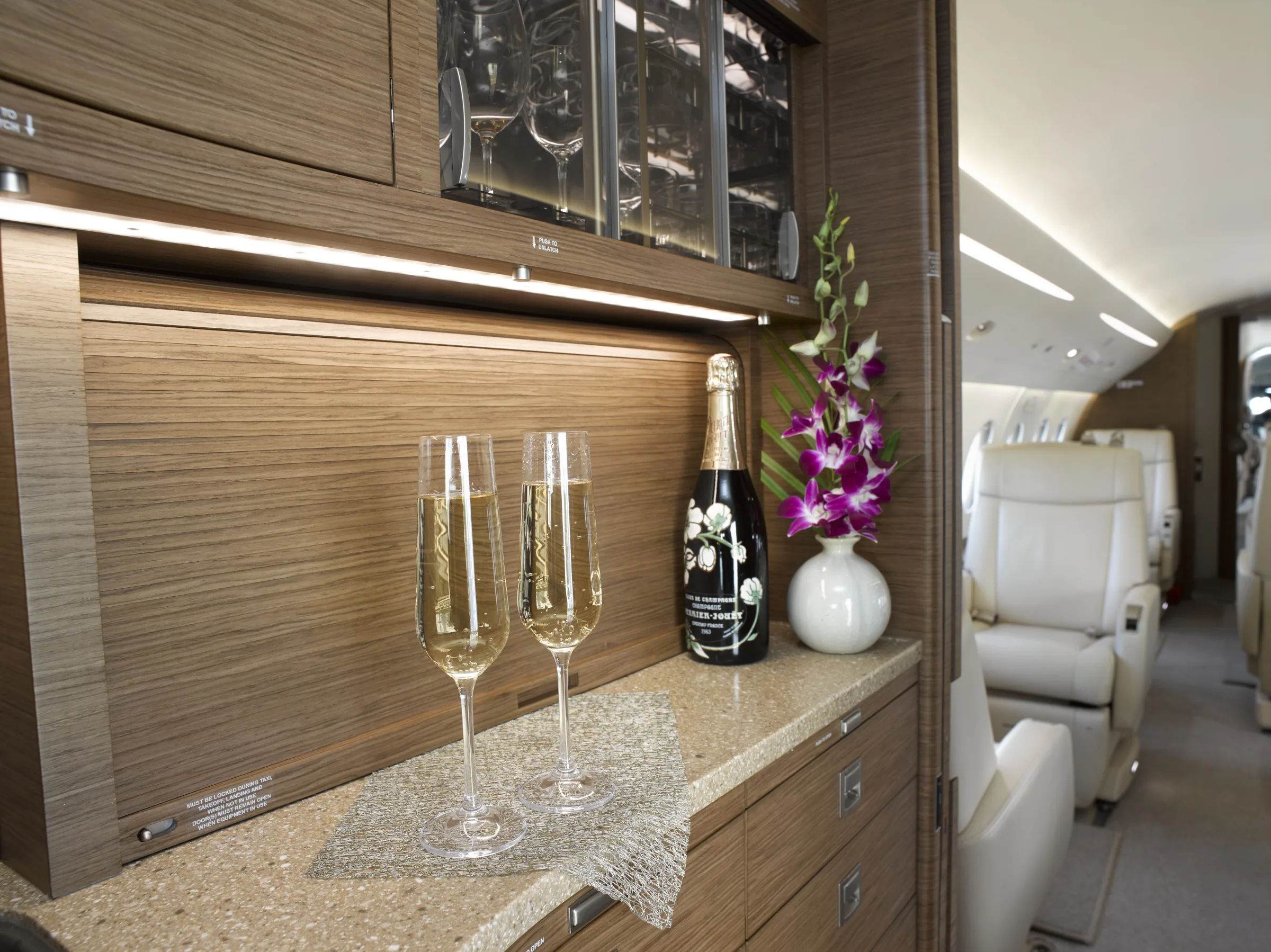 An aircraft galley area with a champagne bottle, two flutes, and purple orchids, with the cabin visible in the background.