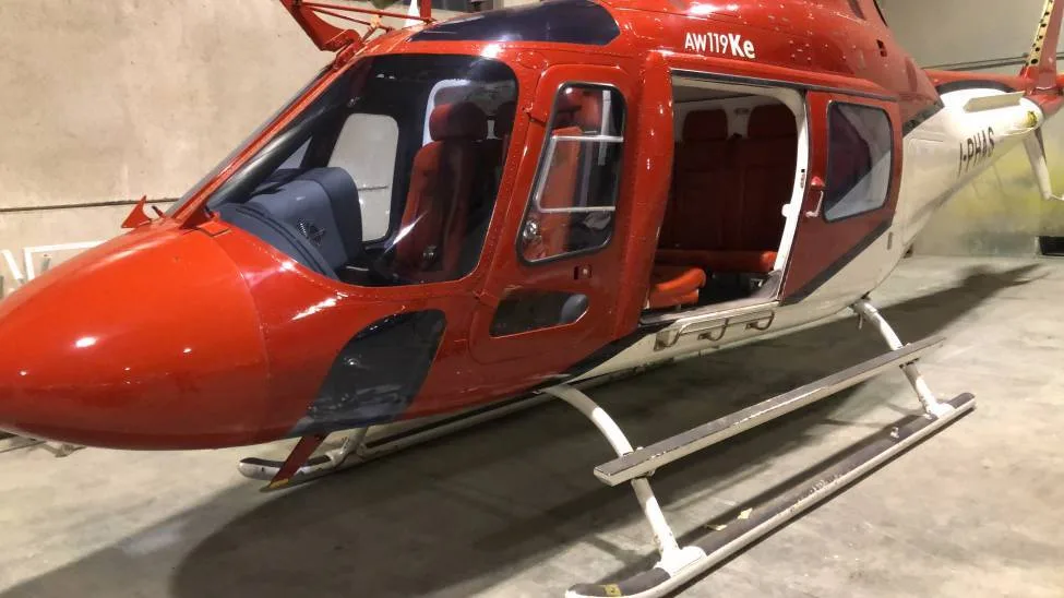 A close-up view of the front and side of a red and white AW119Ke helicopter in a hangar.