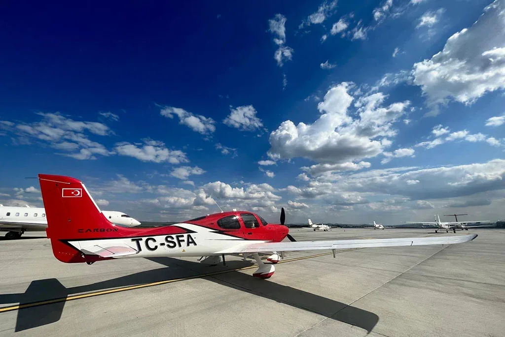 A rear-side view of a red and white single-engine aircraft (TC-SFA) parked on an airfield with other planes in the background.
