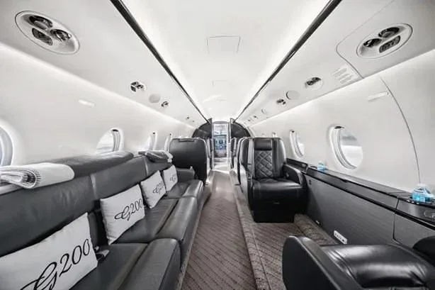 The elegant interior of a private jet cabin, featuring a long black leather sofa, individual seats, and pillows embroidered with 'G200'.