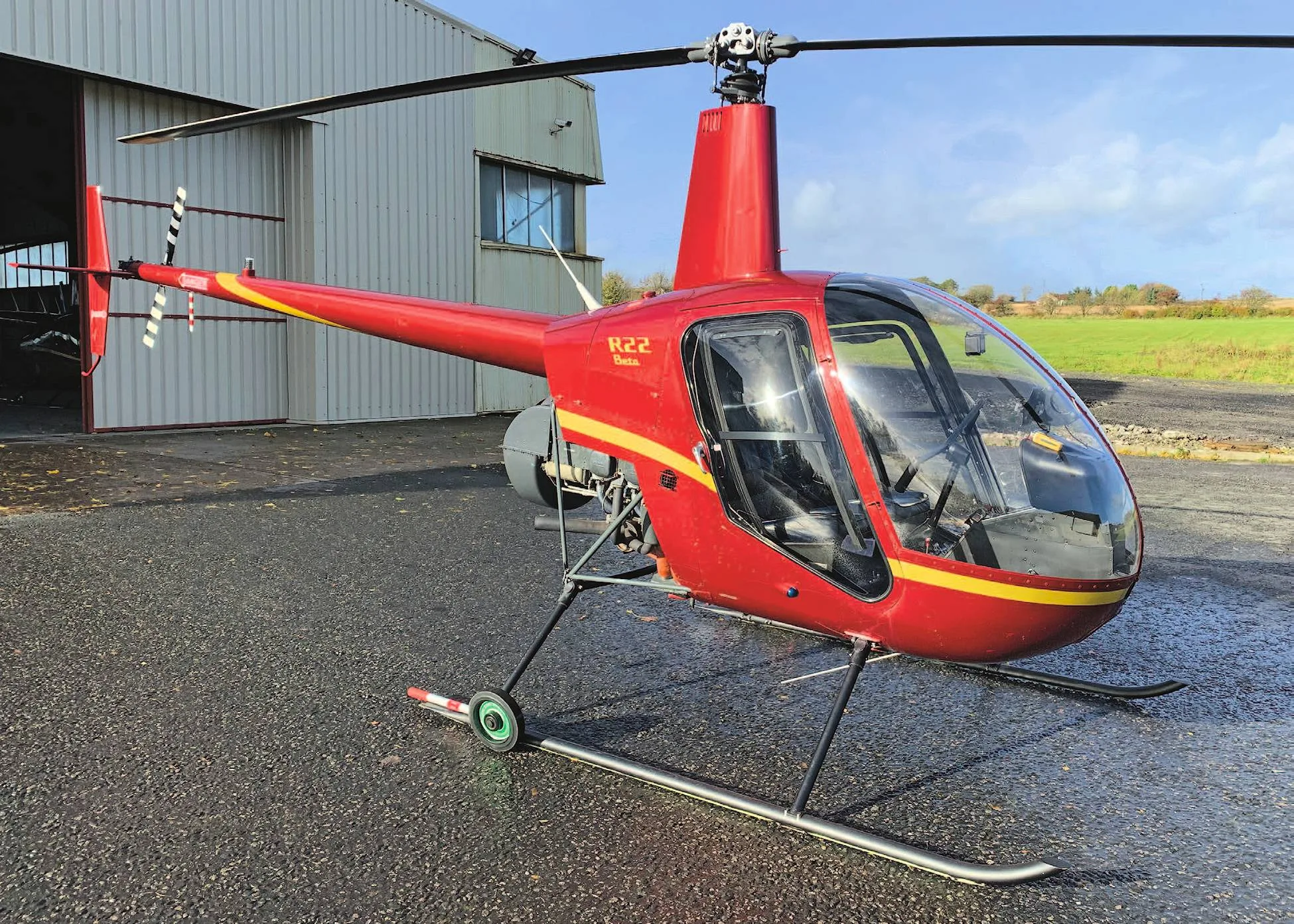 A red Robinson R22 Beta helicopter with yellow stripes parked on a wet tarmac outside a hangar.