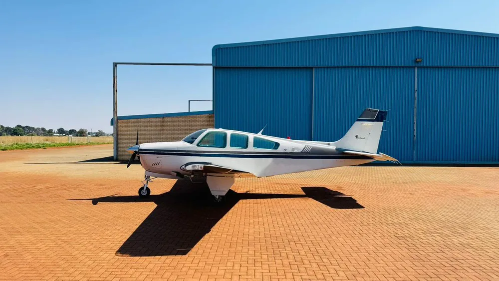 A side view of a white and dark grey single-engine propeller aircraft parked next to a blue hangar.