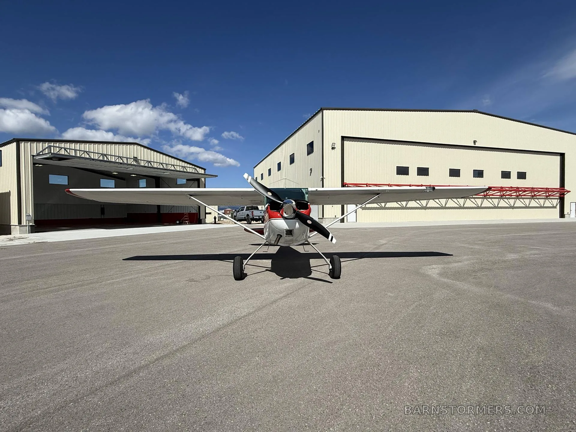 A red and white single-engine propeller aircraft is parked head-on on an asphalt apron in front of two hangars under a blue sky.