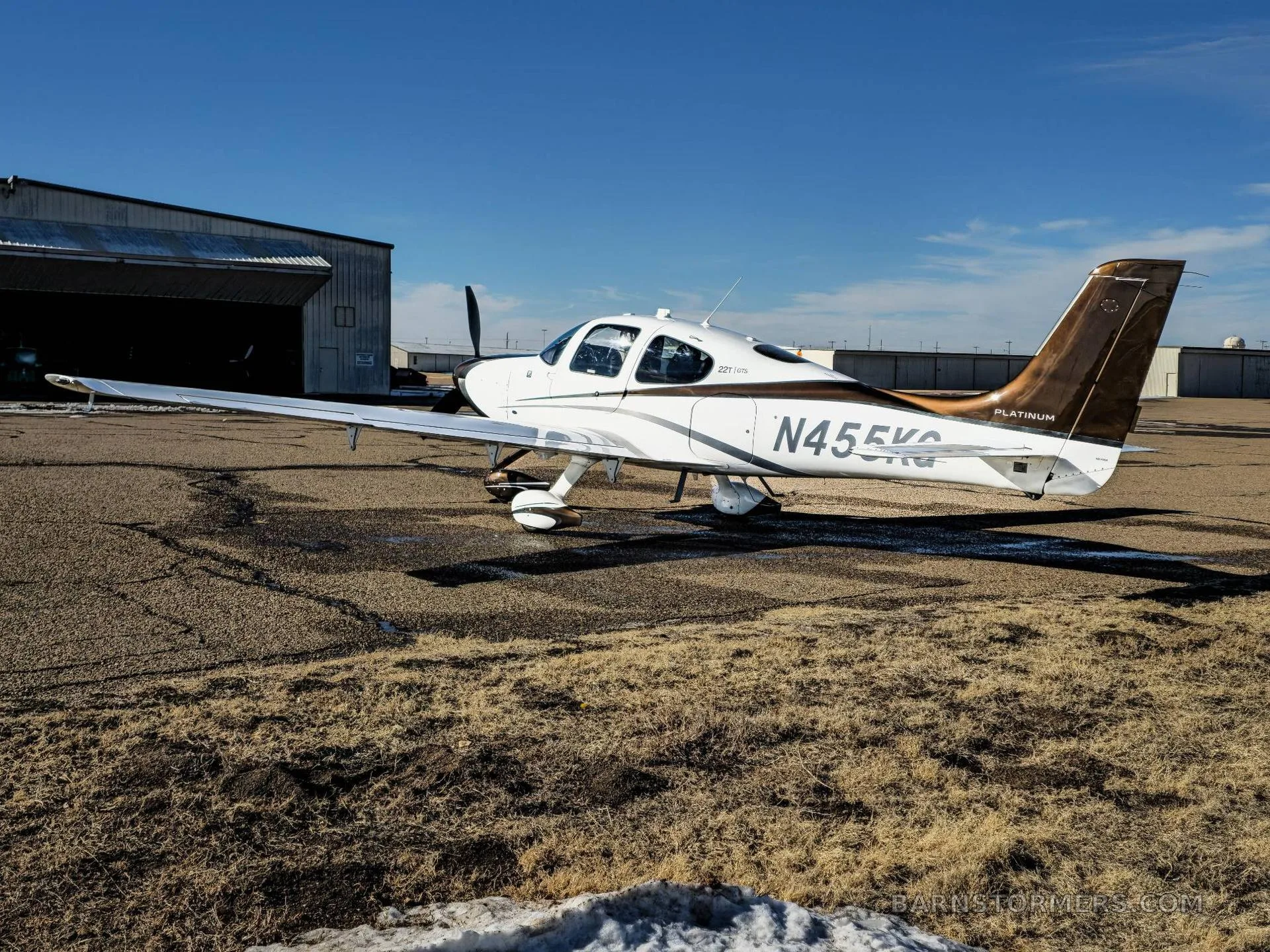 A white and bronze Cirrus SR22T Platinum aircraft (N455KG) parked on an airfield next to a hangar.