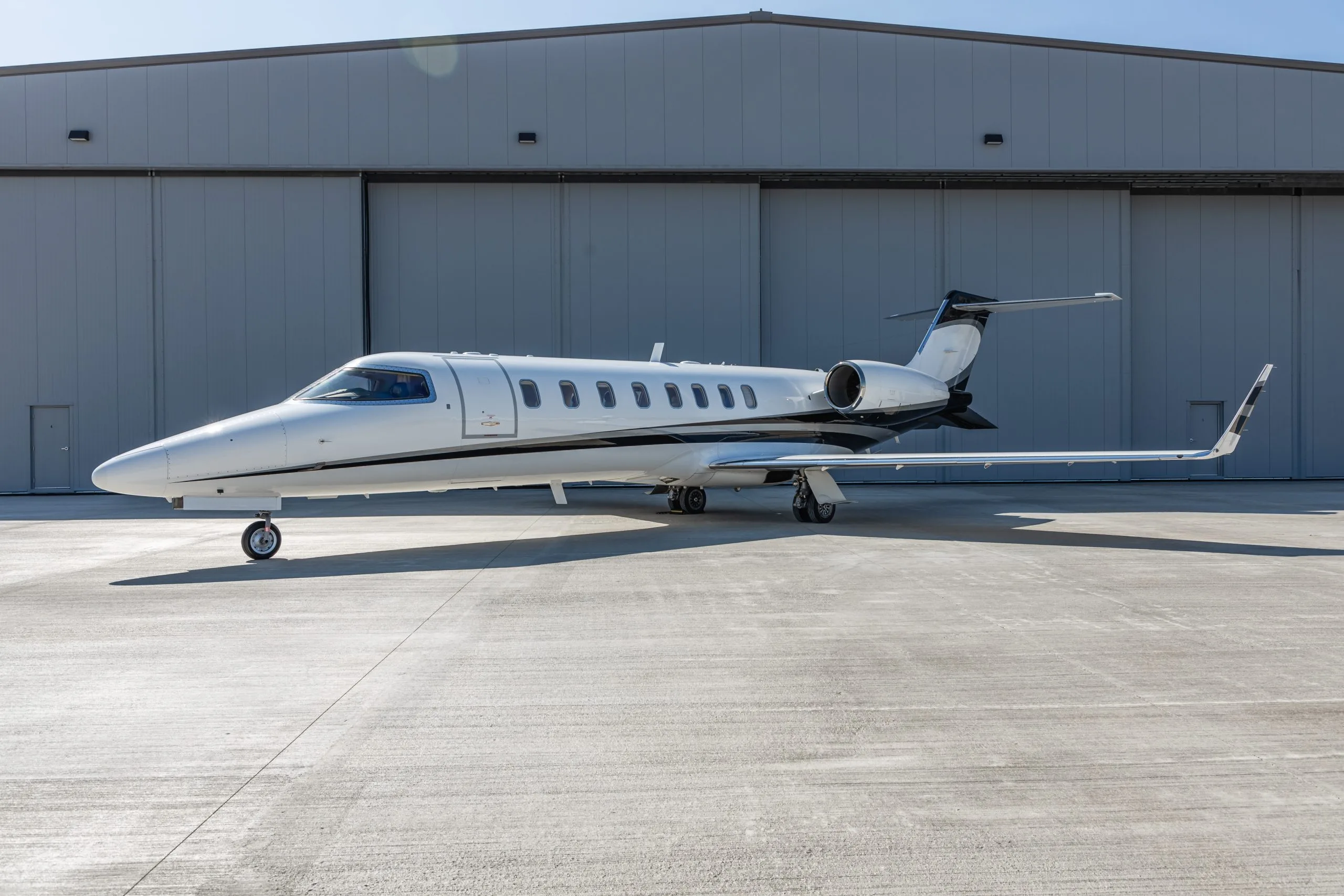 A white business jet with black accents is parked on a tarmac in front of a gray hangar under a clear sky.