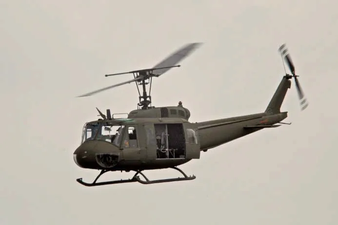An olive drab military helicopter, likely a Bell UH-1 Huey, in flight with its side door open against a light sky.