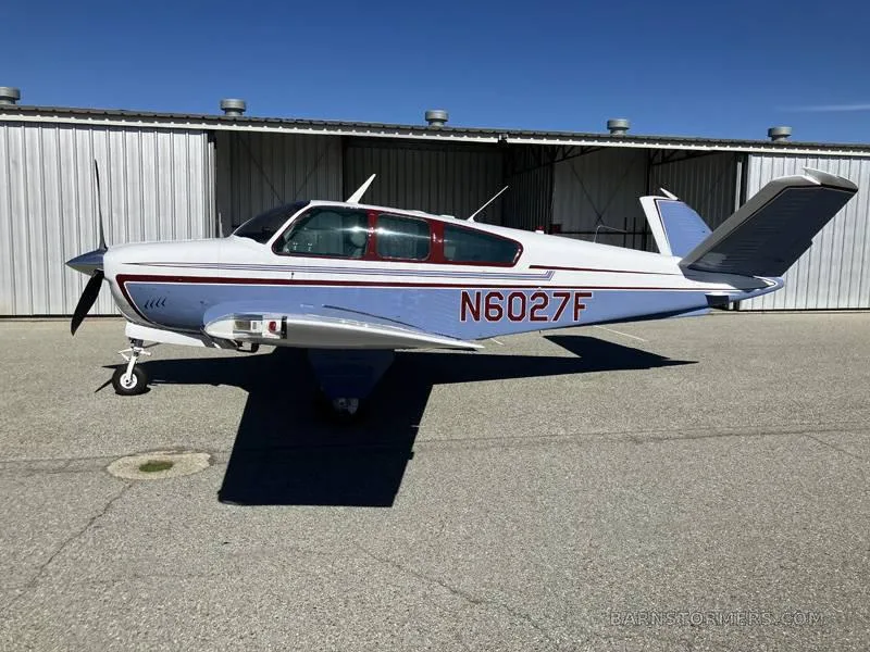 A light blue and white single-engine aircraft with registration N6027F is parked on an asphalt tarmac next to hangars.