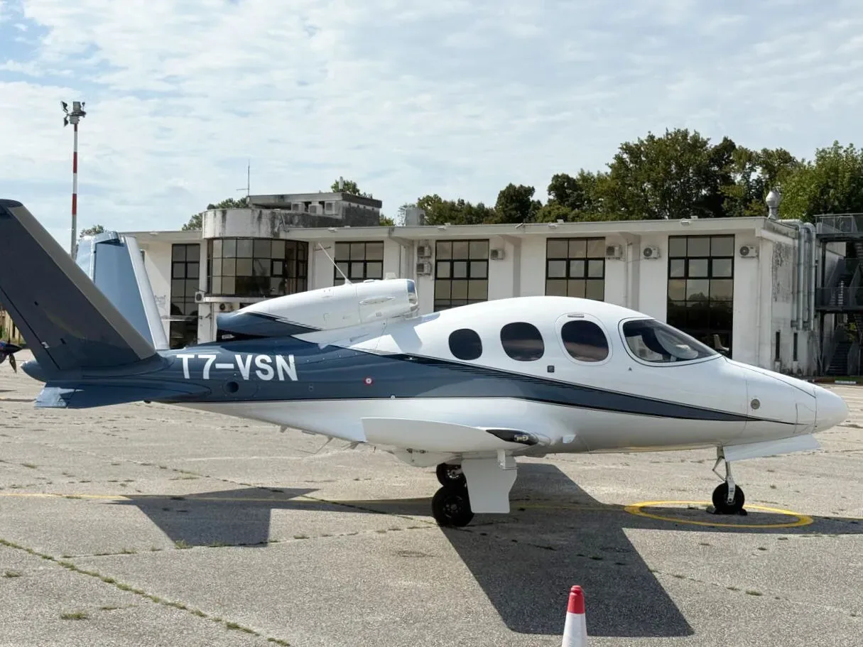 A white and dark blue Cirrus Vision Jet (T7-VSN) parked on an asphalt apron with airport buildings in the background.