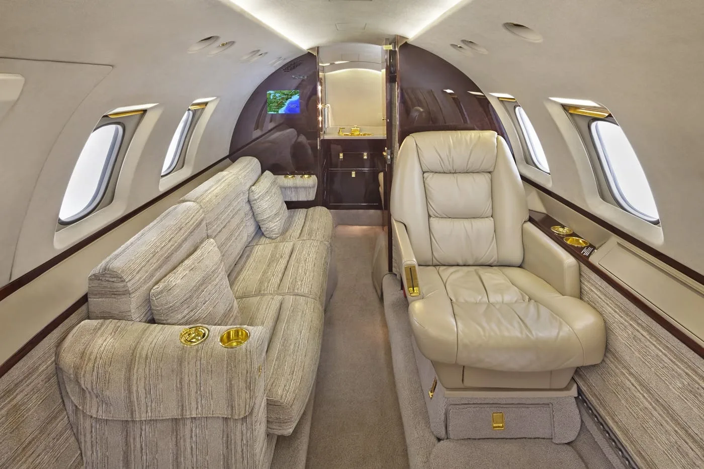 The luxurious interior of a private jet cabin, featuring a beige sofa and a single cream leather seat, with a galley/lavatory area in the background.