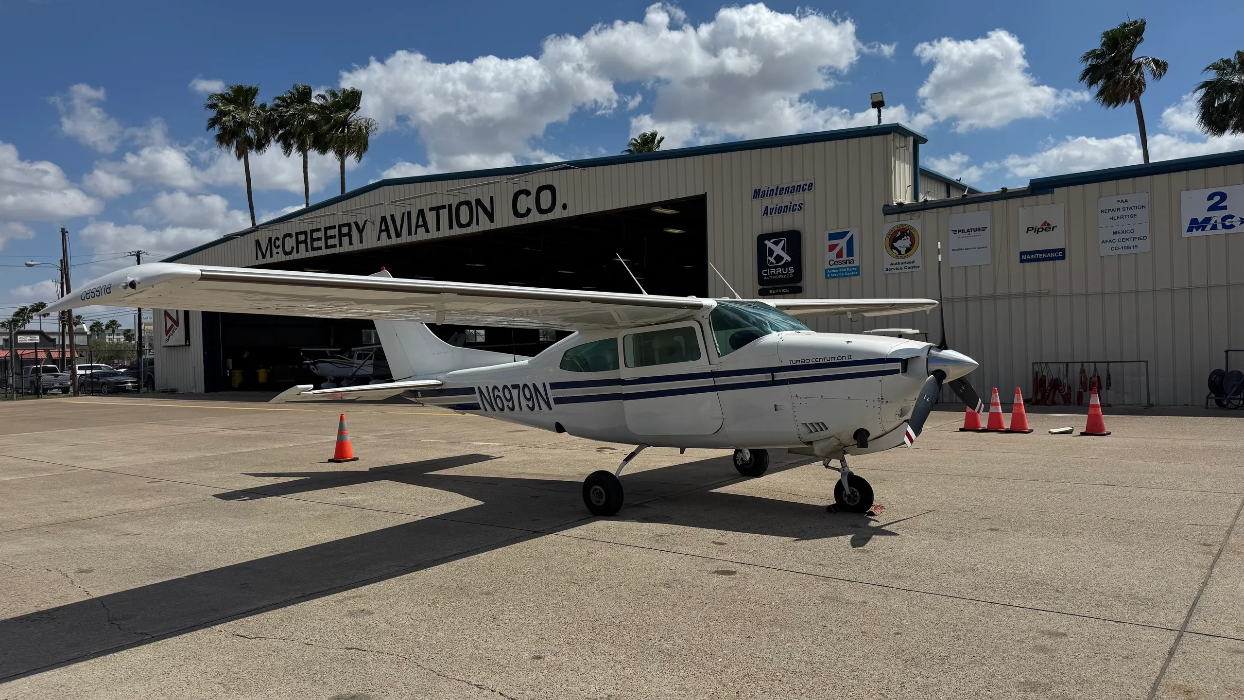 A white Cessna 182 with blue stripes, registration N6979N, parked on a tarmac in front of the McCreery Aviation Co. hangar.