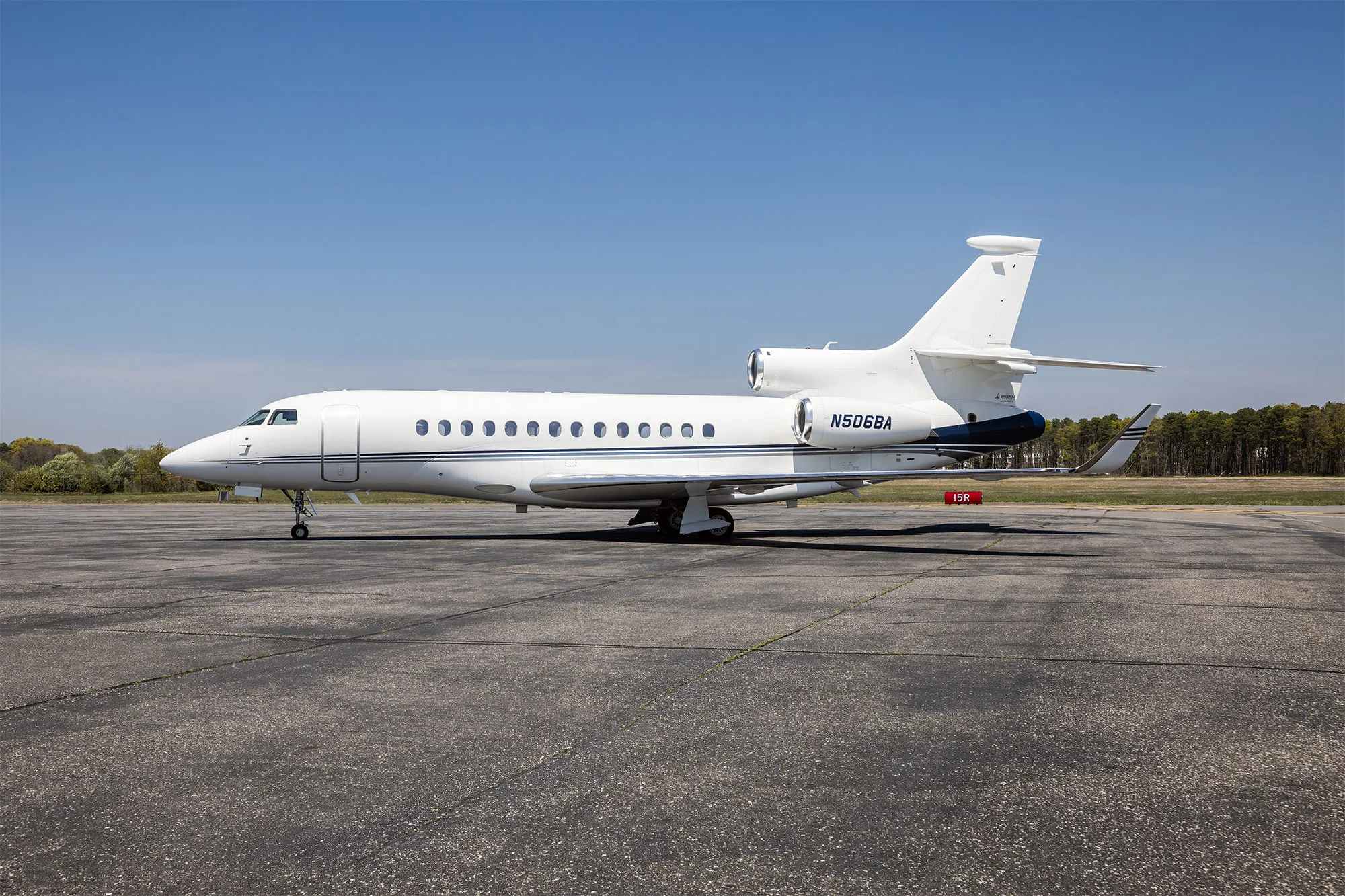A side view of a white Dassault Falcon 900EX business jet with blue accents on the tail, parked on an asphalt tarmac.