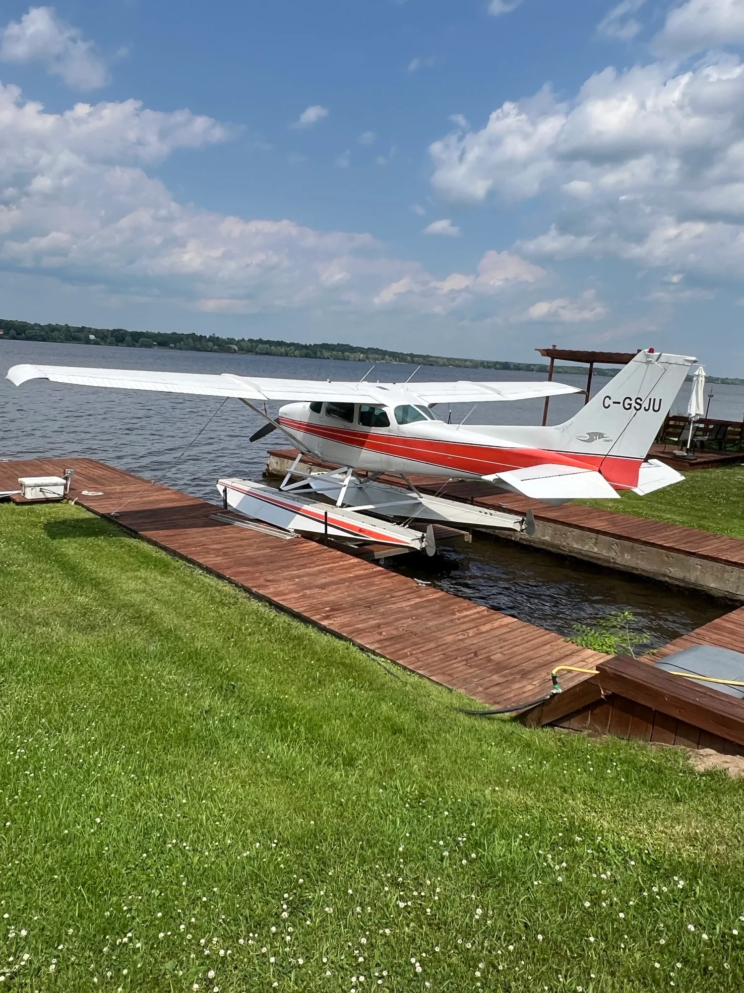 A white and red Cessna Cutlass II floatplane (C-GSJU) is seen from a distance on a wooden dock by a lake.