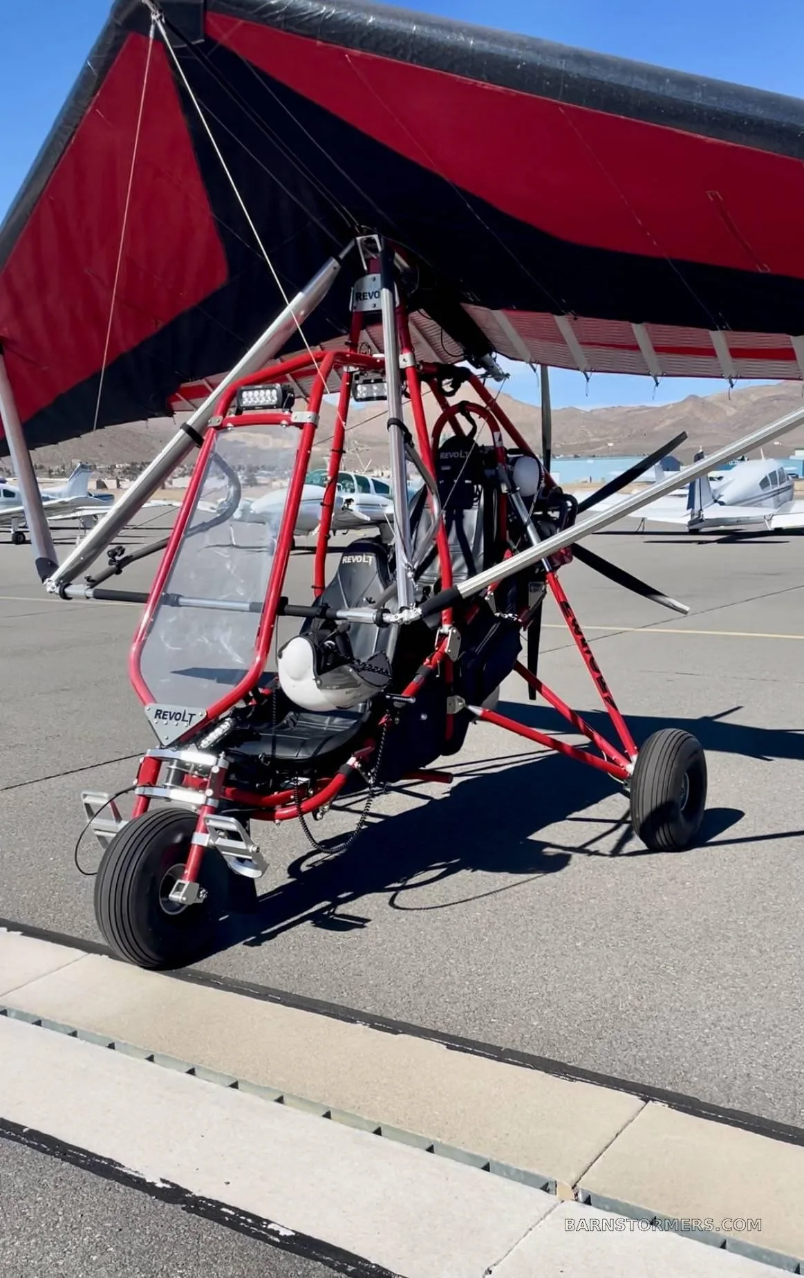 A red and black trike ultralight aircraft with a 'REVOLT' logo on its fairing, parked on an airport tarmac with other aircraft in the background.