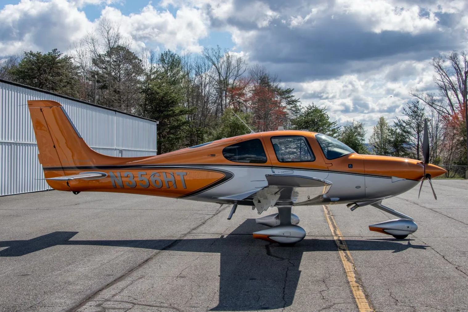 A side view of a metallic orange and silver Cirrus SR22 aircraft (N356HT) parked on an asphalt ramp with trees in the background.