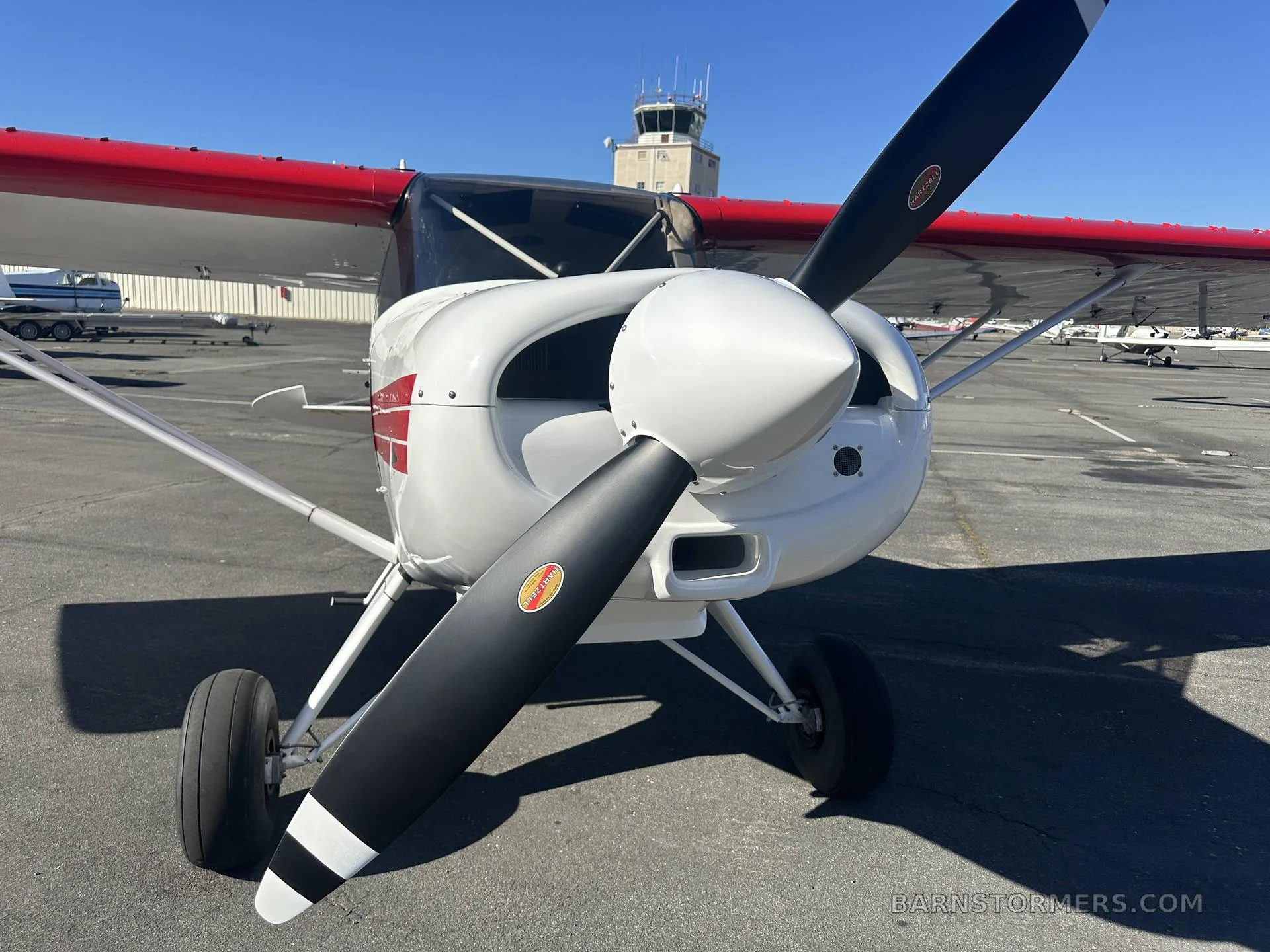 A close-up front view of a white Maule M-7-235 aircraft with a black propeller, parked on an airfield with an ATC tower in the background.